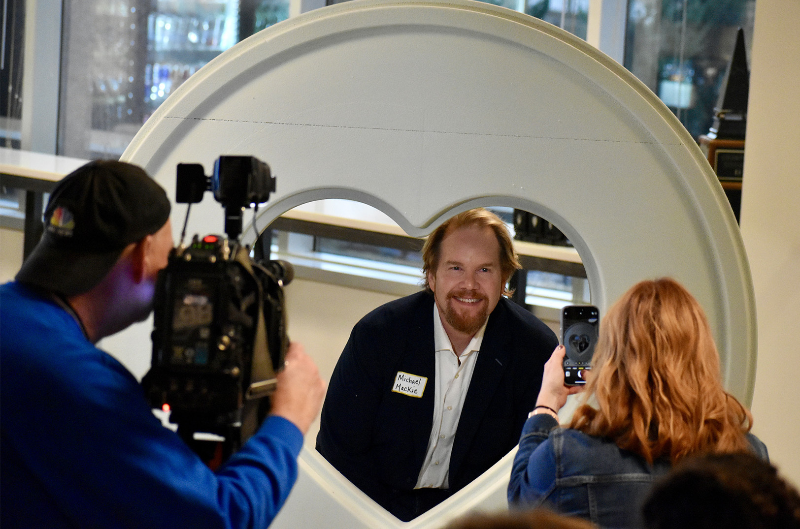Photographers snap images of Michael Mackie, media personality and entertainment reporter, as he poses with the 2026 Parade of Hearts sculpture during a reveal event at nbkc bank; photo by Nikki Overfelt Chifalu, Startland News